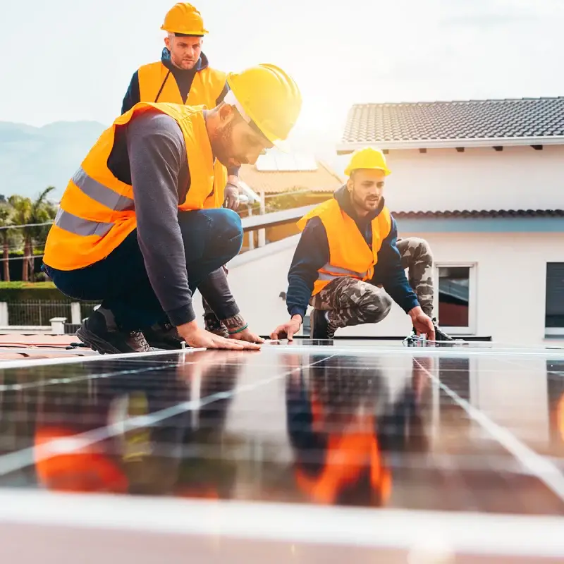 Arbeiter installieren Solarpaneele auf einem Dach bei Sonnenuntergang, tragen Sicherheitswesten und Helme.