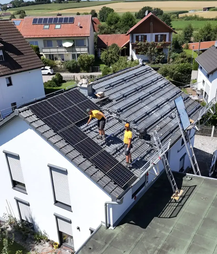 Zwei Arbeiter installieren Solarpanels auf einem Hausdach in einer ländlichen Gegend, um erneuerbare Energie zu nutzen.