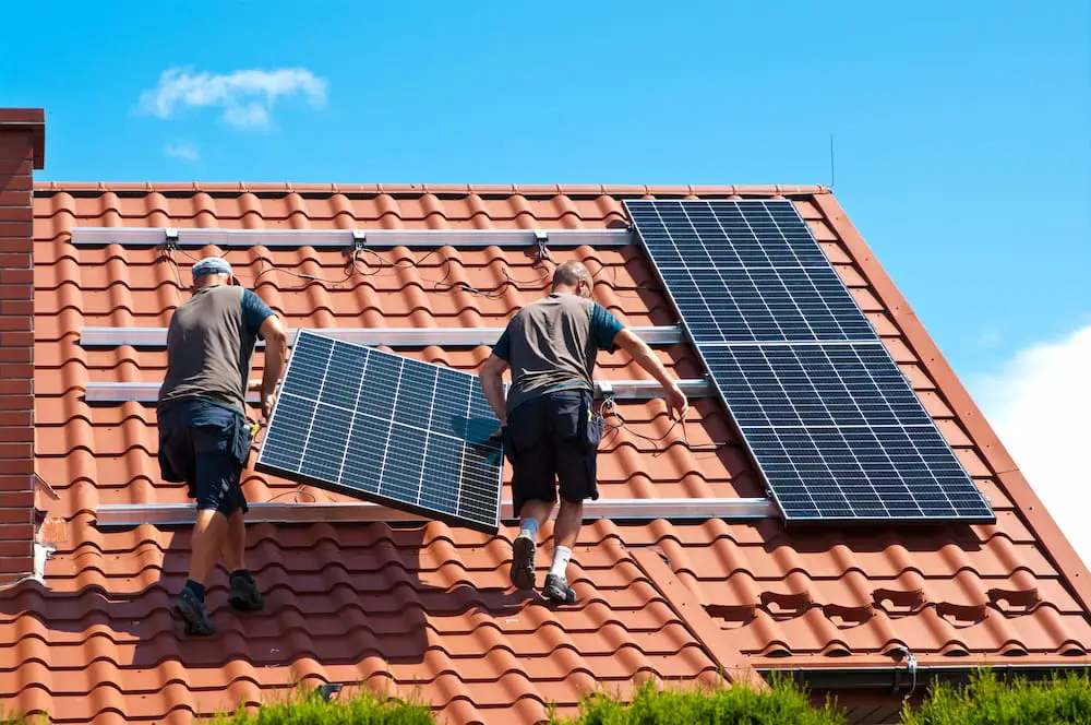 Arbeiter installieren Solarmodule auf einem Ziegeldach unter blauem Himmel.