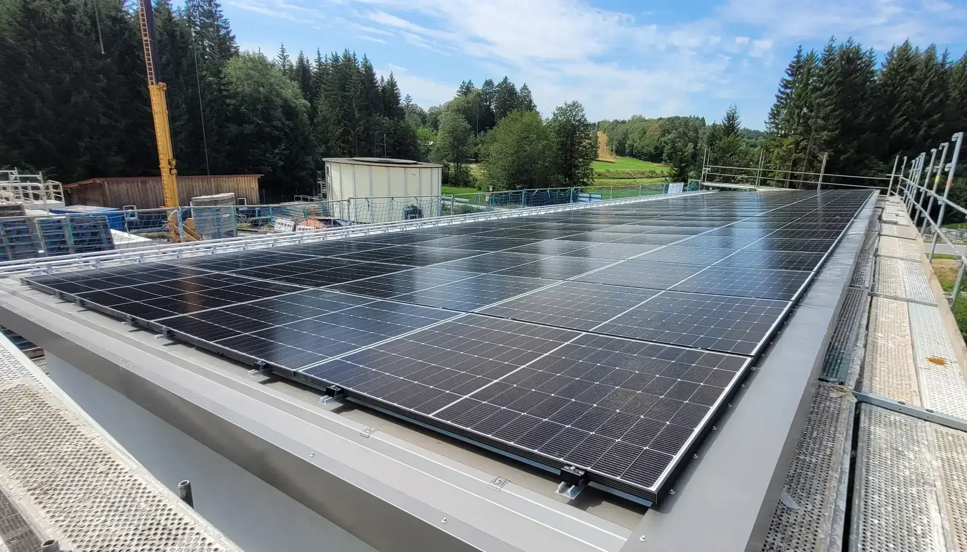 Solaranlage auf dem Dach eines Gebäudes, umgeben von Waldlandschaft unter blauem Himmel.