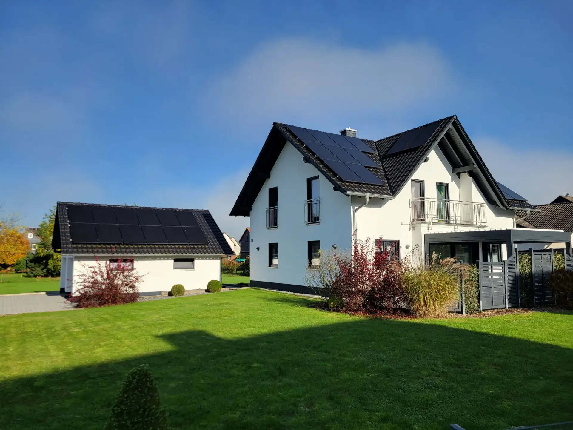 Einfamilienhaus mit Solarpanelen auf Dach und Garage, umgeben von grünem Rasen und blühenden Sträuchern unter blauem Himmel.