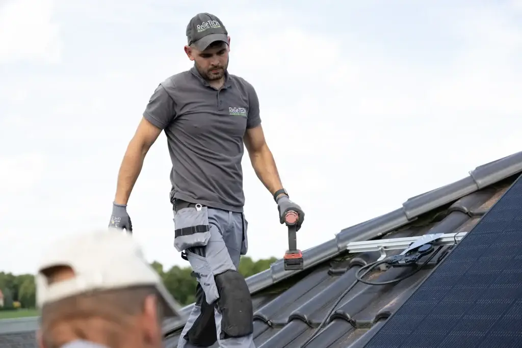Handwerker installiert Solarpanel auf einem Hausdach, trägt Arbeitshose und hält Bohrer. Nachhaltige Energieumrüstung.