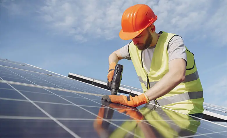 Arbeiter installiert Solarpaneel unter blauem Himmel, trägt Helm und Weste, nutzt Bohrer für Befestigung.
