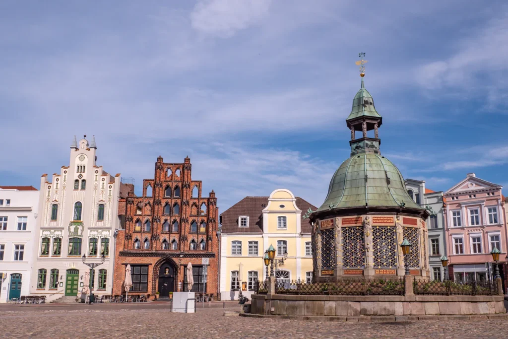 Historischer Marktplatz in Wismar mit Brunnen und farbenfrohen Giebelhäusern bei klarem Himmel.