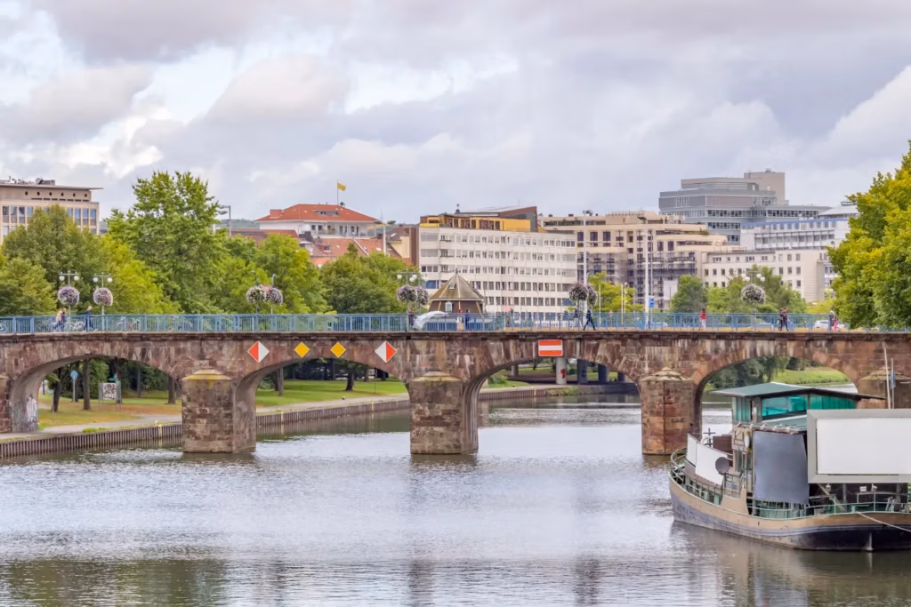 Brücke und Gebäude in Saarbrücken an einem bewölkten Tag, mit einem Boot auf der Saar im Vordergrund.