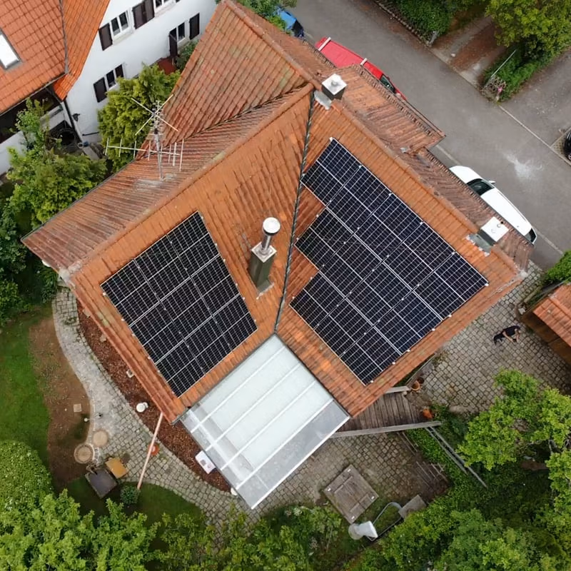 Drohnenansicht eines Hauses mit Solaranlagen auf dem Dach, umgeben von Garten und Einfahrt in einer Wohngegend.